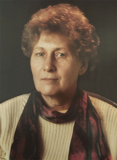 Portr&auml;t einer &auml;lteren Frau mit lockigem Haar, einem hellen Pullover und einem gepunkteten Schal. Der Hintergrund ist dunkel.