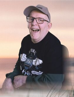 L&auml;chelnder Mann mit Brille und Baseballkappe, bekleidet mit T-Shirt in grauen und schwarzen T&ouml;nen, vor einem hellen Hintergrund.