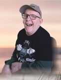 L&auml;chelnder Mann mit Brille und Baseballkappe, bekleidet mit T-Shirt in grauen und schwarzen T&ouml;nen, vor einem hellen Hintergrund.