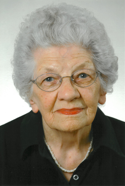 Portr&auml;t einer &auml;lteren Frau mit grauen Locken, Brille und rotem Lippenstift, bekleidet mit einem schwarzen Oberteil.