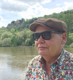 Person mit Sonnenbrille und farbenfrohem Hemd steht am Fluss, im Hintergrund eine Burg auf einem H&uuml;gel.