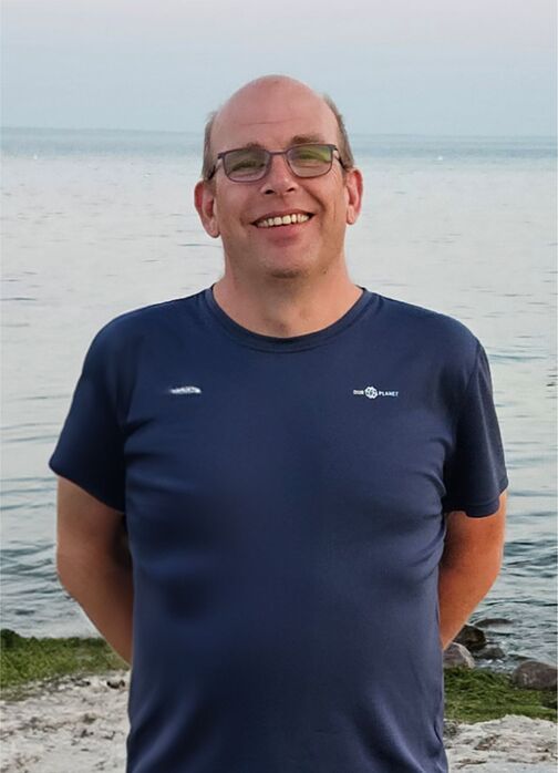 L&auml;chelnder Mann mit Brille in dunkelblauem Shirt steht am Wasser, im Hintergrund ruhige Wasseroberfl&auml;che und flacher Strand.