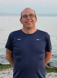L&auml;chelnder Mann mit Brille in dunkelblauem Shirt steht am Wasser, im Hintergrund ruhige Wasseroberfl&auml;che und flacher Strand.