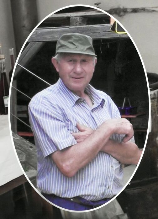 &Auml;lterer Mann mit einem gr&uuml;nen Hut und gestreiftem Hemd, stehend mit verschr&auml;nkten Armen vor einer Werkstatt.