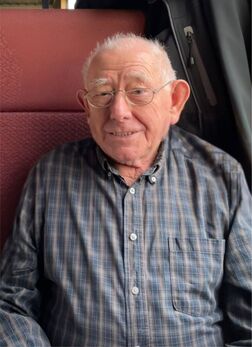 &Auml;lterer Mann mit Brille und kariertem Hemd sitzt l&auml;chelnd auf einer roten Sitzbank.