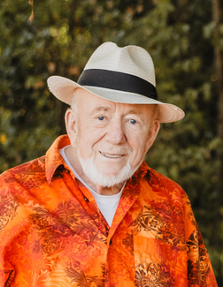 L&auml;chelnder &auml;lterer Mann mit Bart, Hut und buntem, floralem Hemd vor unscharfem, gr&uuml;nem Hintergrund.