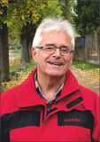 L&auml;chelnder Mann in roter Jacke steht auf einem herbstlichen Weg, umgeben von B&auml;umen und fallenden Bl&auml;ttern.