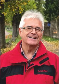 L&auml;chelnder Mann in roter Jacke steht auf einem herbstlichen Weg, umgeben von B&auml;umen und fallenden Bl&auml;ttern.