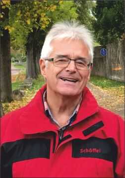 L&auml;chelnder Mann in roter Jacke steht auf einem herbstlichen Weg, umgeben von B&auml;umen und fallenden Bl&auml;ttern.