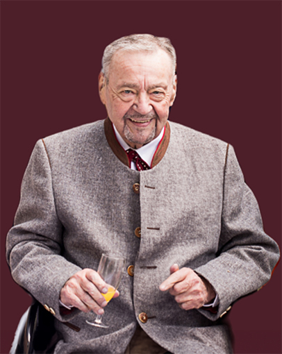 &Auml;lterer Mann in traditioneller Tracht mit einem Sektglas sitzt l&auml;chelnd vor einem dunkelroten Hintergrund.