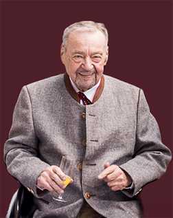 &Auml;lterer Mann in traditioneller Tracht mit einem Sektglas sitzt l&auml;chelnd vor einem dunkelroten Hintergrund.