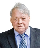 Mann mit grauem Anzug und kariertem Blau-Weiß-Tie steht vor neutralem Hintergrund und blickt direkt in die Kamera.