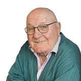 Lächelnder älterer Mann mit Brille und grünem Pullover, vor neutralem Hintergrund.