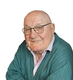 L&auml;chelnder &auml;lterer Mann mit Brille und gr&uuml;nem Pullover, vor neutralem Hintergrund.