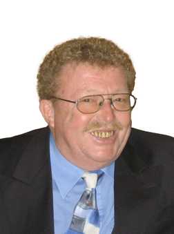 L&auml;chelnder Mann mit lockigem Haar, Brille und Anzug, tr&auml;gt eine gestreifte Krawatte und hat einen freundlichen Ausdruck.