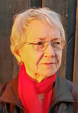 Portr&auml;t einer &auml;lteren Frau mit kurzen, grauen Haaren und Brille, die einen roten Schal und eine schwarze Jacke tr&auml;gt.