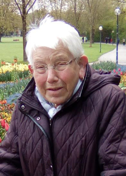 &Auml;ltere Frau mit Brille und hellem Haar, l&auml;chelt vor bunten Blumen im Park.