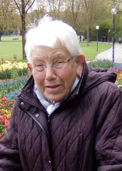 &Auml;ltere Frau mit Brille und hellem Haar, l&auml;chelt vor bunten Blumen im Park.