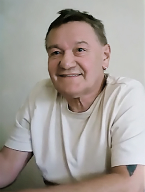 L&auml;chelnder Mann in einem wei&szlig;en T-Shirt sitzt an einem Tisch vor hellem Hintergrund.