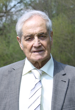 Portr&auml;t eines &auml;lteren Mannes in einem Anzug mit gestreifter Krawatte, vor einem unscharfen, gr&uuml;nen Hintergrund.