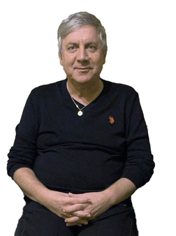 Mann mit grauen Haaren sitzt in einem schwarzen Pullover und h&auml;lt die H&auml;nde auf dem Scho&szlig;. Hintergrund ist wei&szlig;.