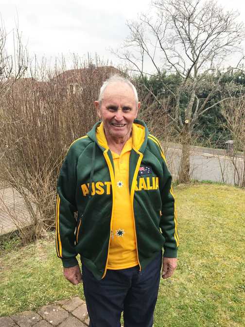 &Auml;lterer Mann in einem gr&uuml;nen Australien-Sweatshirt und gelbem Shirt steht im Garten mit Str&auml;uchern im Hintergrund.