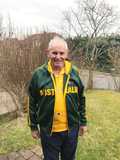 &Auml;lterer Mann in einem gr&uuml;nen Australien-Sweatshirt und gelbem Shirt steht im Garten mit Str&auml;uchern im Hintergrund.