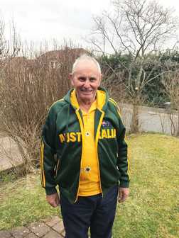 &Auml;lterer Mann in einem gr&uuml;nen Australien-Sweatshirt und gelbem Shirt steht im Garten mit Str&auml;uchern im Hintergrund.