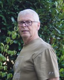 Mann mit grauen Haaren und Brille steht vor grünen Pflanzen, trägt ein olivfarbenes T-Shirt mit Aufdruck.