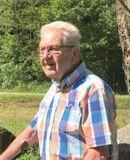 &Auml;lterer Mann mit Brille steht vor einer gr&uuml;nen Landschaft und tr&auml;gt ein karierter Hemd in Blau-Orange-T&ouml;nen.