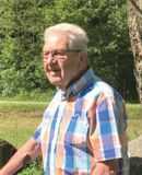 &Auml;lterer Mann mit Brille steht vor einer gr&uuml;nen Landschaft und tr&auml;gt ein karierter Hemd in Blau-Orange-T&ouml;nen.