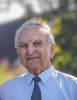 &Auml;lterer Mann mit grauen Haaren, blauem Hemd und Krawatte, l&auml;chelt freundlich vor unscharfem Hintergrund.