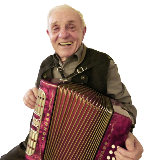 L&auml;chelnder &auml;lterer Mann mit einem roten Akkordeon in der Hand, tr&auml;gt eine schwarze Weste und ein graues Hemd.
