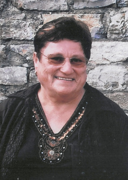 Portr&auml;t einer l&auml;chelnden &auml;lteren Frau mit dunklem Oberteil und Brille vor einer steinernen Wand.