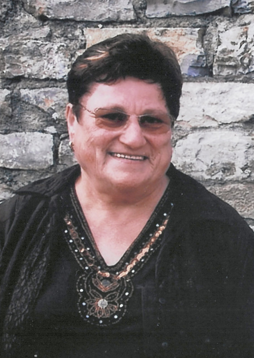 Portr&auml;t einer l&auml;chelnden &auml;lteren Frau mit dunklem Oberteil und Brille vor einer steinernen Wand.