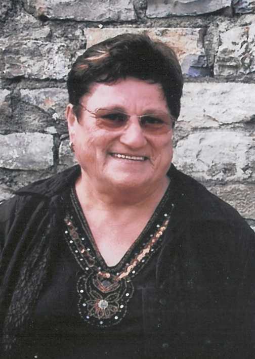 Portr&auml;t einer l&auml;chelnden &auml;lteren Frau mit dunklem Oberteil und Brille vor einer steinernen Wand.