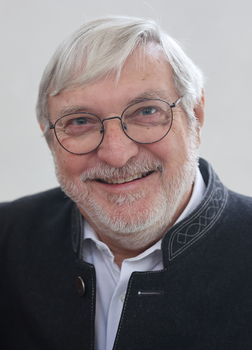 Nahaufnahme eines l&auml;chelnden Mannes mit Brille und grauem Bart, gekleidet in einer dunklen Jacke mit Verzierungen.