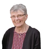 Ältere Frau mit kurzen grauen Haaren, Brille und lächelndem Gesicht, trägt ein gemustertes Oberteil und eine schwarze Jacke.