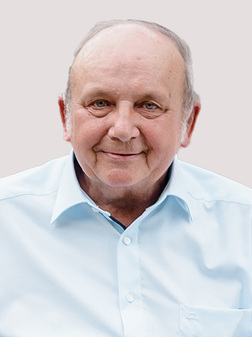 Portrait eines &auml;lteren Mannes mit kurzem, grauem Haar, der in einem hellblauen Hemd l&auml;chelt. Hintergrund ist neutral.