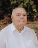 Älterer Mann mit ernster Miene in weißem Hemd, im Hintergrund grüne Blätter.