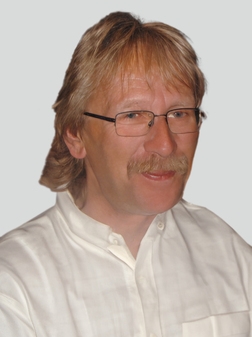 Porträt eines lächelnden Mannes mit Brille und blonden, leicht gewellten Haaren, in einem weißen Hemd.