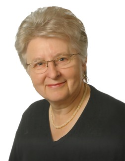 Portrait einer &auml;lteren Frau mit grauen Haaren, Brille und einer Perlenkette, l&auml;chelnd vor einem wei&szlig;en Hintergrund.