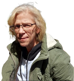 Frau mit kurzen, blonden Haaren und Brille, trägt eine grüne Jacke, schaut direkt in die Kamera.