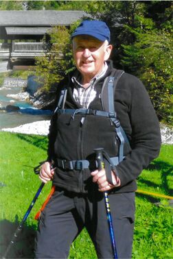 Mann mit Wanderstock in outdoor Kleidung steht vor einer gr&uuml;nen Wiese und einem kleinen Fluss, im Hintergrund eine Holzbr&uuml;cke.