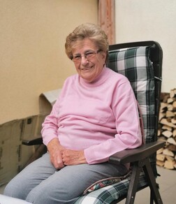 &Auml;ltere Frau in einem Stuhl, l&auml;chelnd, tr&auml;gt einen rosa Pullover und sitzt neben einem Stapel Holzscheite.