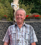 Ein Mann steht auf Steinstufen vor einer Statue von Jesus am Kreuz, umgeben von Bäumen und Blumen.