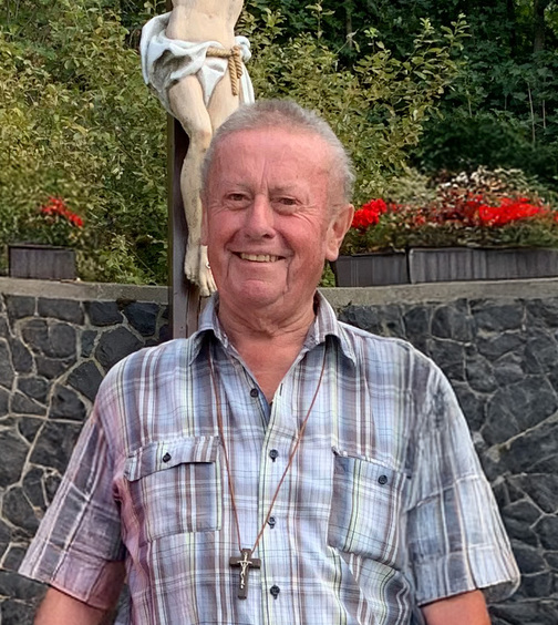 Ein Mann steht auf Steinstufen vor einer Statue von Jesus am Kreuz, umgeben von Bäumen und Blumen.