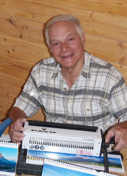 Ein &auml;lterer Mann l&auml;chelt, w&auml;hrend er eine Spiralbindemaschine vor sich h&auml;lt, umgeben von Kalenderbl&auml;ttern.