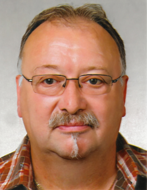 Portr&auml;t eines mittelalten Mannes mit Brille, Halbglatze und Schnurrbart, in kariertem Hemd vor grauem Hintergrund.