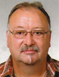 Porträt eines mittelalten Mannes mit Brille, Halbglatze und Schnurrbart, in kariertem Hemd vor grauem Hintergrund.